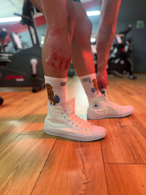 Person wearing white sneakers and socks with a brand logo on a wooden floor.